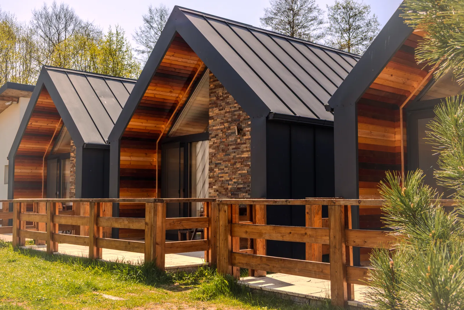 Trei cabane moderne cu acoperiș negru, placare de lemn și piatră, cu gard de lemn în față