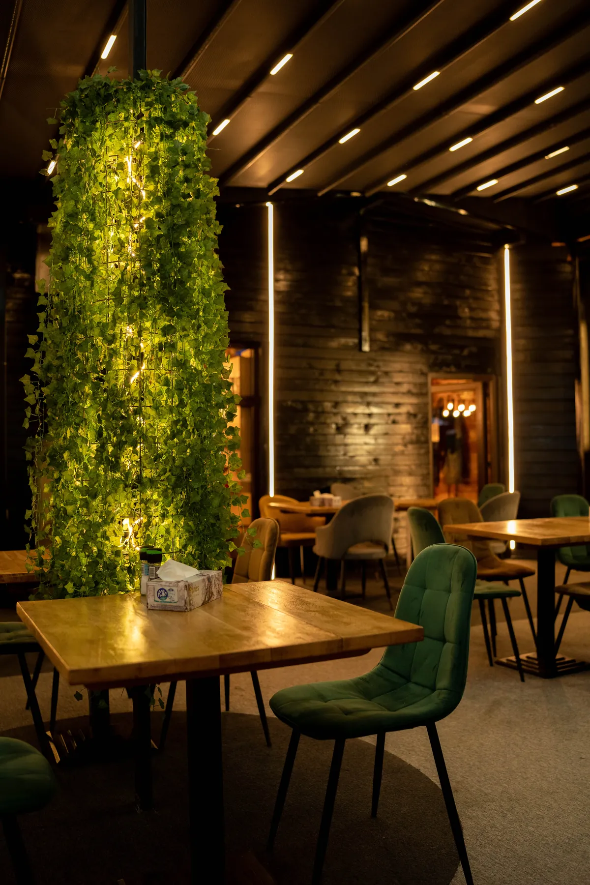 Interiorul restaurantului seara: un stâlp îmbrăcat în iederă luminată, mese de lemn și scaune de catifea verde