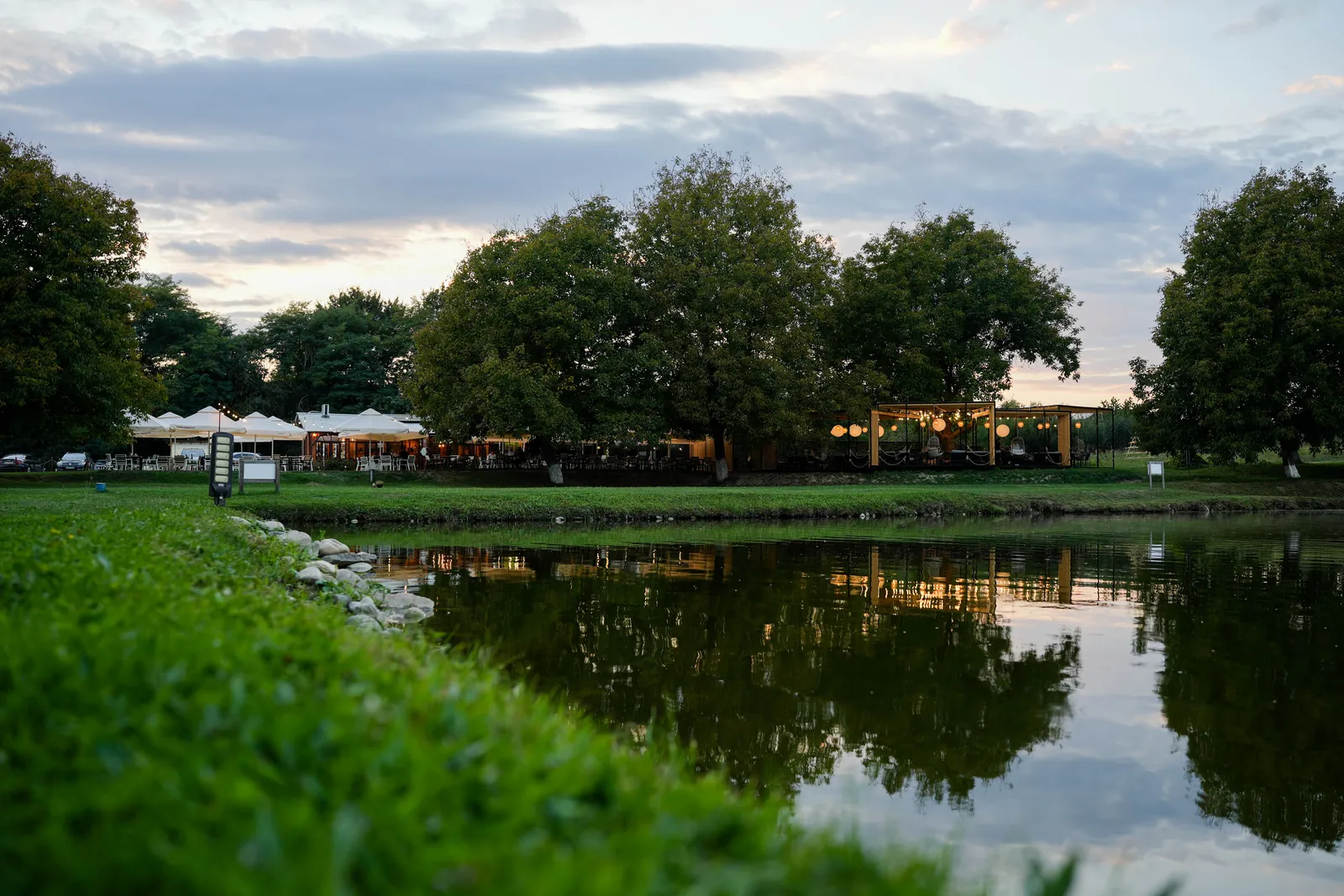 Restaurant pe malul unui lac, la apus, cu terasa luminată reflectată în apă